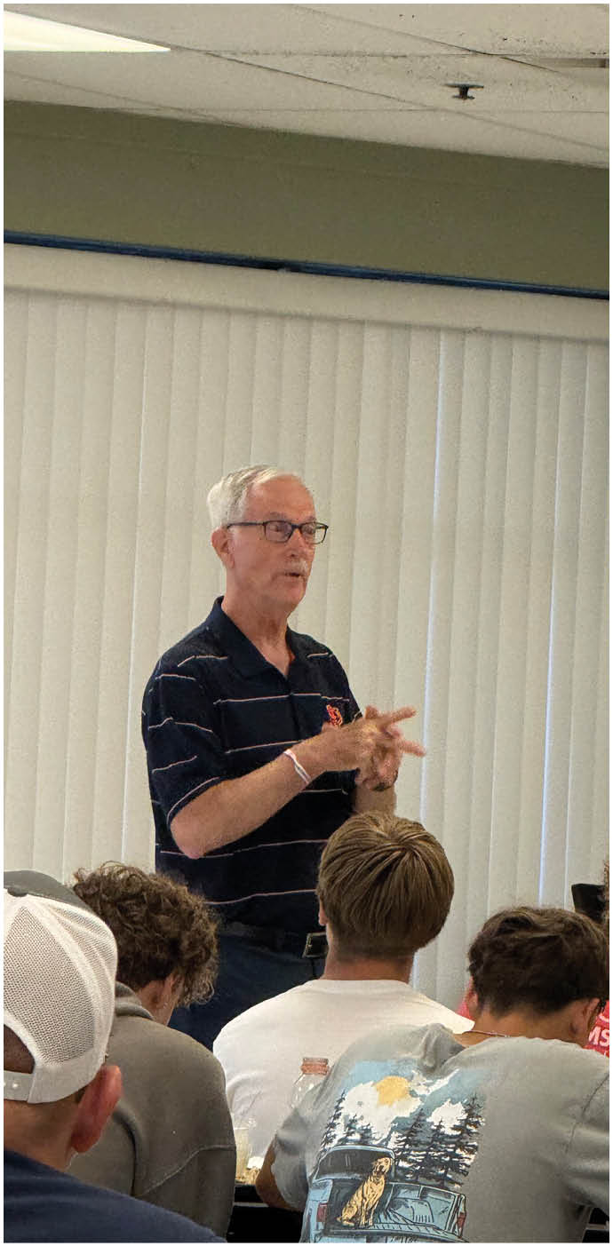 A senior person wearing glasses and a striped polo shirt is speaking and gesturing in front of a group of seated people in a room with vertical blinds. Description generated by AI