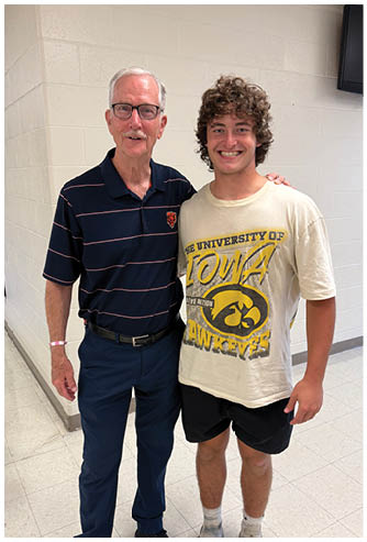 Two people stand side by side smiling in a hallway, one wearing a striped polo shirt and glasses and the other wearing a University of Iowa Hawkeyes t-shirt and shorts. Description generated by AI