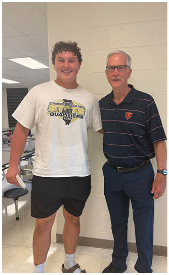 Two people stand side by side indoors with one wearing a white t-shirt and black shorts and the other wearing a dark striped polo shirt and dark pants. Description generated by AI