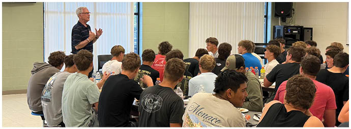 A senior person wearing glasses and a striped shirt is standing and speaking to a group of seated people in a classroom setting with large windows and blinds. Description generated by AI