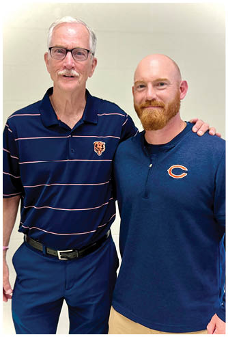 Two adults stand close together wearing dark blue tops, one with a striped polo shirt featuring a bear logo and the other with a quarter-zip pullover. Description generated by AI