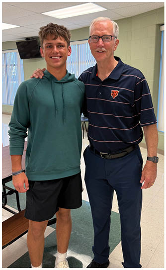 Two people stand side by side indoors with one person wearing a green hoodie and black shorts and the other wearing a navy blue striped polo shirt and dark pants. Description generated by AI