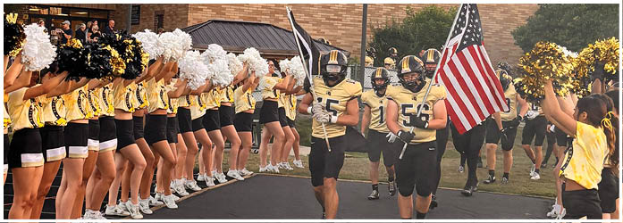 A group of people in yellow and black uniforms are holding pom-poms and cheering while others in football gear hold flags, including an American flag, in front of a brick building. Description generated by AI