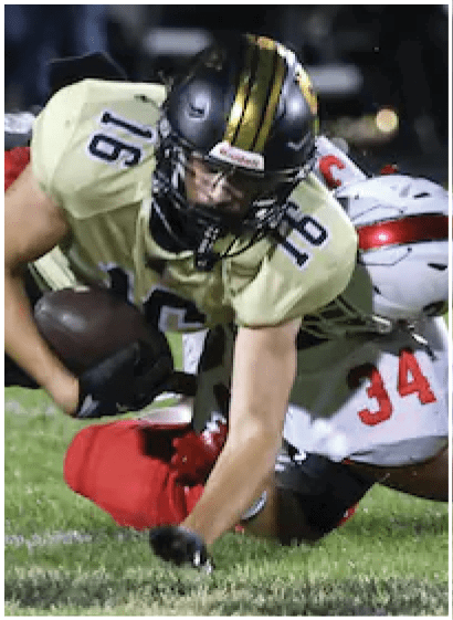A person wearing a football uniform with the number 16 is diving forward while holding a football, with another player in red attempting to tackle them on a grassy field. Description generated by AI