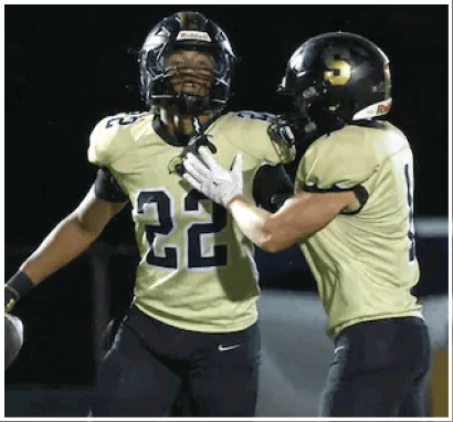 Two people wearing football helmets and uniforms celebrate on a field at night with one holding a football. Description generated by AI
