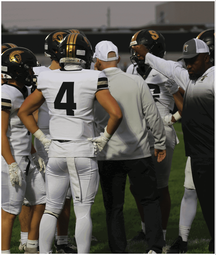 A group of people in football uniforms and coaches are gathered on a field during a game or practice. Description generated by AI