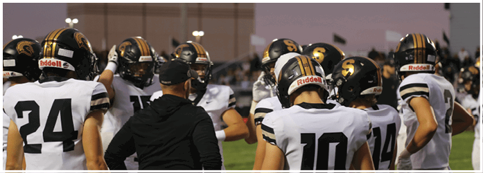 A group of people wearing football helmets and white jerseys with black numbers are gathered on a field, seemingly in a team huddle or discussion. Description generated by AI