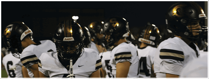 A group of people wearing white football uniforms and black helmets stand closely together on a field at night. Description generated by AI