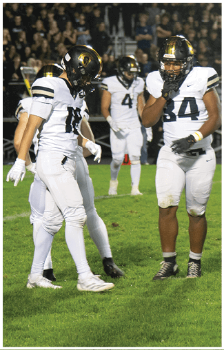 Three people in white football uniforms and helmets stand on a grassy field at night with a crowd in the background. Description generated by AI