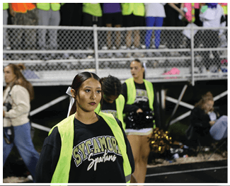 A person wearing a Sycamore Spartans shirt and a neon yellow vest stands on a track field with other people and bleachers in the background. Description generated by AI