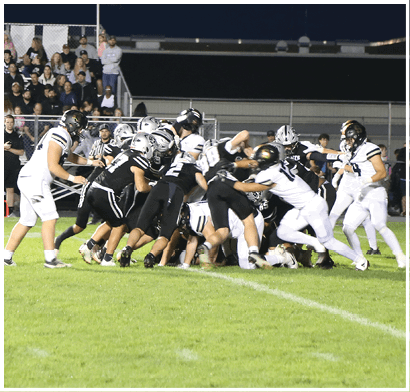 A group of people in football uniforms are engaged in a play on a grassy field at night with spectators watching from bleachers in the background. Description generated by AI