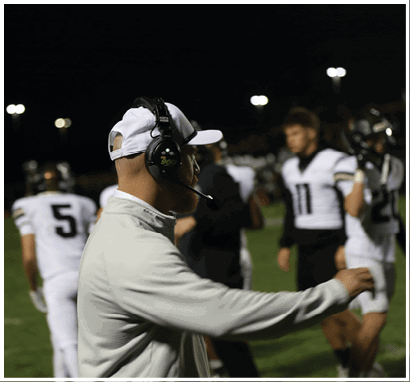 A person wearing a white cap and headset is gesturing on a sports field at night while several people in white and black sports uniforms stand in the background. Description generated by AI