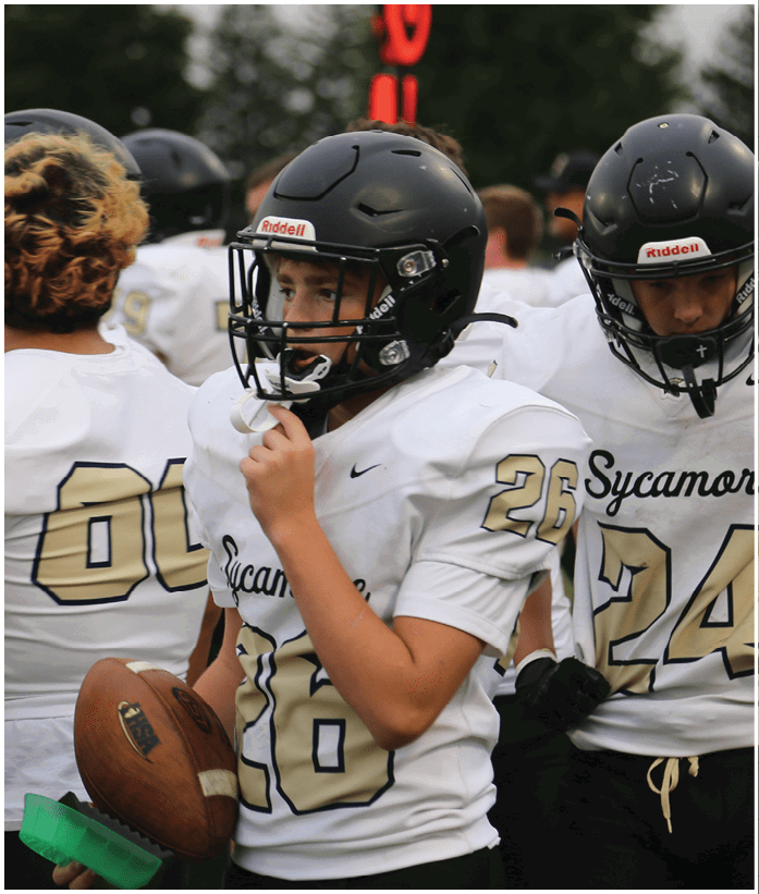People wearing football helmets and white jerseys are gathered on a field, with one person holding a football and adjusting their helmet. Description generated by AI