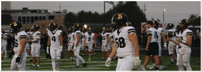 People wearing white football uniforms with black helmets are standing and walking on a football field during dusk. Description generated by AI