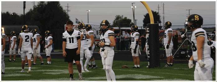 People in football uniforms are standing and walking on a field near a goalpost during what appears to be a practice or warm-up session. Description generated by AI