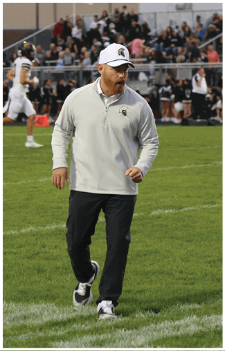 A person wearing a white cap and light gray pullover is walking on a grassy sports field with players and spectators in the background. Description generated by AI