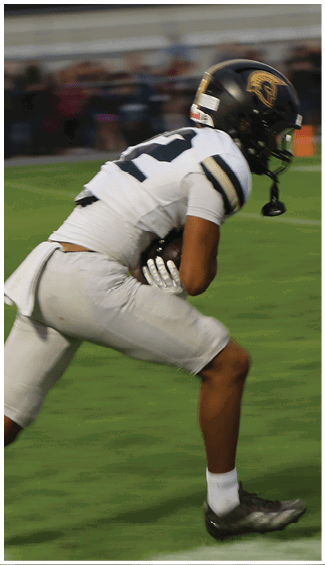 A person wearing a white football uniform and black helmet is running on a grassy field while holding a football. Description generated by AI