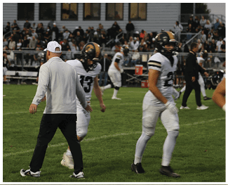 People in football uniforms and a person in casual sportswear are on a grassy field with spectators in the background. Description generated by AI