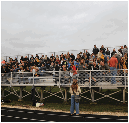 A person stands on a track facing a crowd of people seated on bleachers under a cloudy sky. Description generated by AI