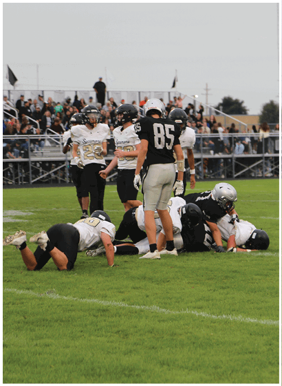 People wearing football uniforms are engaged in a play on a grassy field with spectators in the background. Description generated by AI