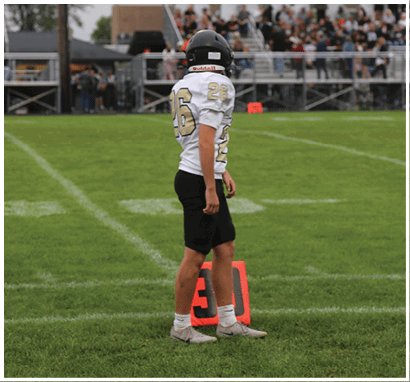 A person wearing a football uniform with the number 26 stands on a grassy field near an orange yard marker with spectators in the background. Description generated by AI