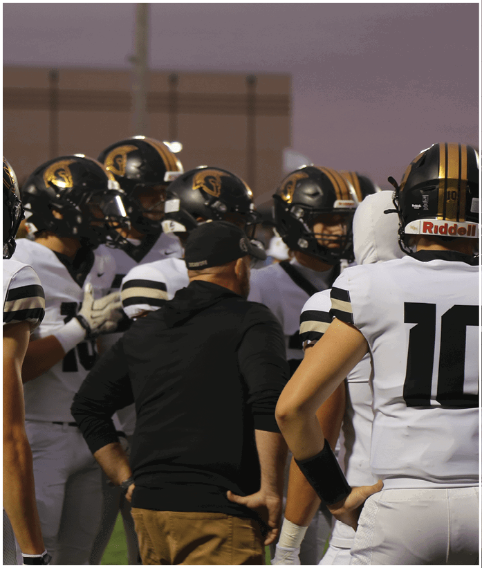 A group of people wearing football helmets and uniforms are gathered closely together on a field with one person in a black jacket appearing to give instructions. Description generated by AI