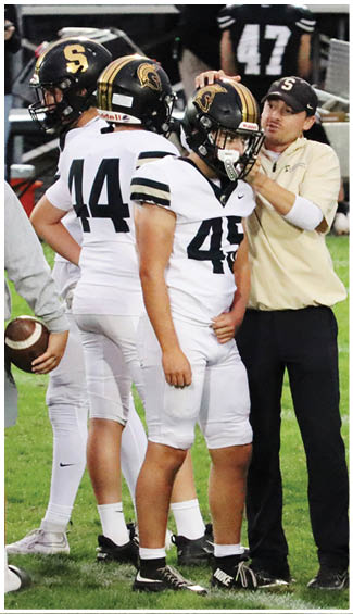 A person in a white football uniform with the number 43 stands next to another person in a beige shirt and black pants on a grassy field. Description generated by AI