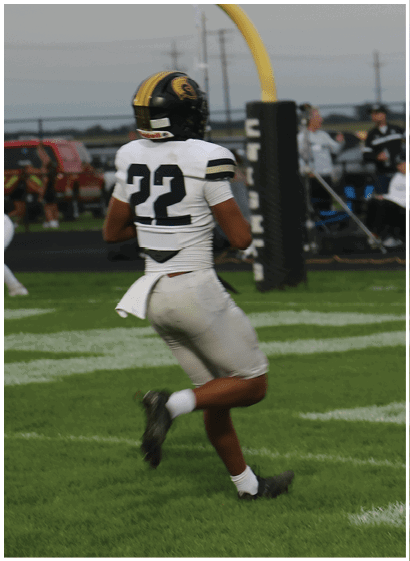 A person wearing a white football uniform with the number 22 is running on a grassy field toward a yellow goalpost with spectators and vehicles in the background. Description generated by AI