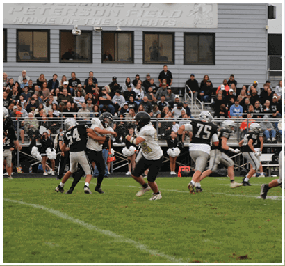 A group of people in football uniforms are playing on a grassy field with a crowd of spectators watching from bleachers in front of a building labeled “Peter J. Noll Home of the Knights." Description generated by AI