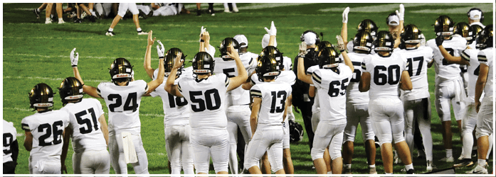 A group of people wearing football uniforms and helmets stand on a field with their arms raised, some pointing upwards. Description generated by AI