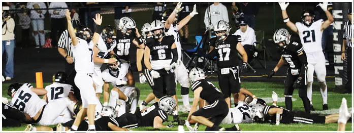 Players in football uniforms and helmets are raising their hands on a field, with some kneeling and others standing, indicating a moment of celebration or a call during a game. Description generated by AI