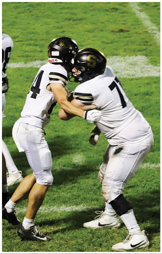 Two people in white football uniforms and black helmets are engaged in a blocking drill on a grassy field. Description generated by AI