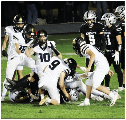 Several people in football uniforms are gathered on a grassy field with some standing and others on the ground during a game. Description generated by AI