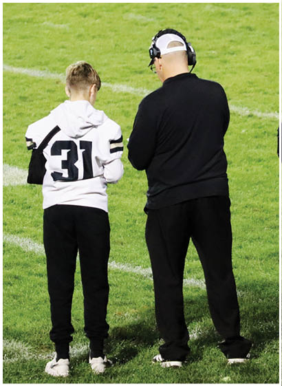 Two people stand on grass, one wearing a white hoodie with the number 31 and black pants, and the other wearing a black outfit. Description generated by AI
