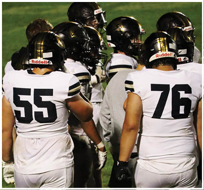People wearing white football uniforms with black helmets are gathered on a field, with two players numbered 55 and 76 prominently visible from the back. Description generated by AI