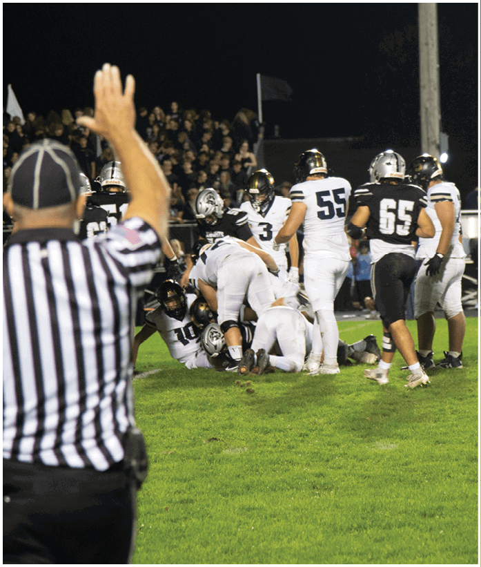 A referee raises one arm signaling during a nighttime football game where players in white and black uniforms are gathered on the field with a crowd in the background. Description generated by AI