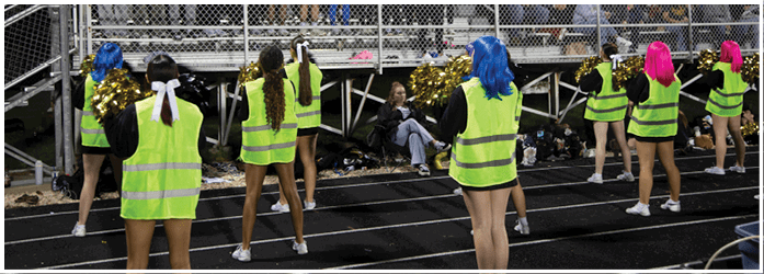 A group of people in yellow and black uniforms are holding pom-poms and cheering while others in football gear hold flags, including an American flag, in front of a brick building. Description generated by AI