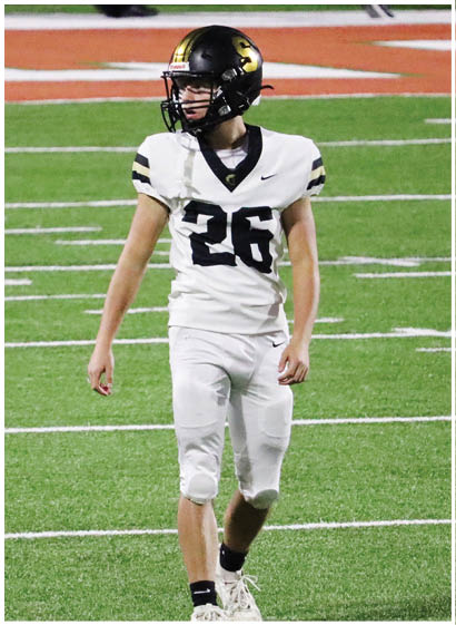 A person wearing white athletic pants and a white shirt is standing on a green sports field with white lines. Description generated by AI