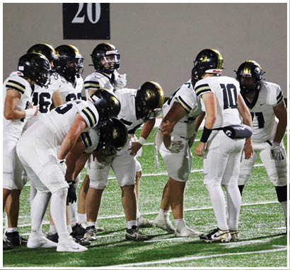 A group of people wearing white football uniforms and black helmets are huddled together on a green field. Description generated by AI