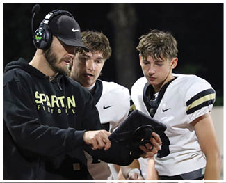 An adult wearing a headset and cap is showing a tablet to two young athletes in white football uniforms during a nighttime outdoor event. Description generated by AI