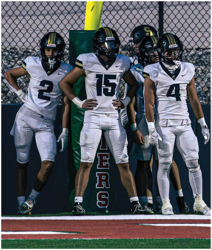 Three people wearing white football uniforms stand on a field near a goalpost with the word “TIGERS" visible in the background. Description generated by AI