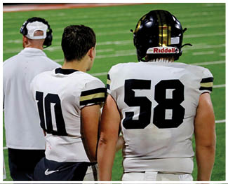 Two people wearing white football uniforms with numbers 10 and 58 stand on a green field with a third person in a white jacket and cap nearby. Description generated by AI