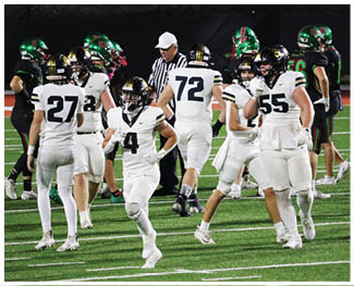 Adults in football uniforms from two teams are on a field with one team wearing white and the other wearing black and green helmets, with a referee in the background. Description generated by AI