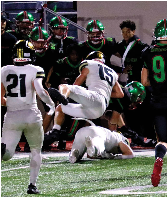 Several people wearing green helmets and dark uniforms are closely watching or engaging with two people in white sports uniforms during an intense moment in a football game. Description generated by AI
