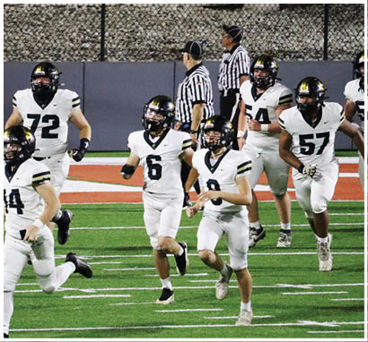 People wearing white football uniforms with black helmets are running on a green field during a game. Description generated by AI