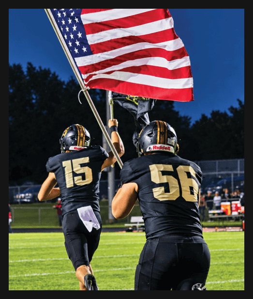 Two people wearing helmets hold up a waving American flag against a dark blue sky. Description generated by AI