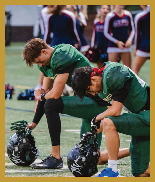 Two people in green football uniforms kneel on a field holding their helmets with heads bowed while cheerleaders stand blurred in the background. Description generated by AI