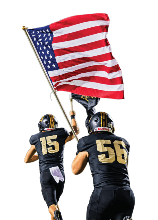 Two people in black football uniforms run on a field with one holding a large American flag against a dark evening sky. Description generated by AI