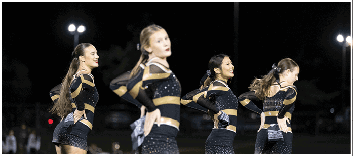 Four adults in matching black and gold dance costumes perform a synchronized routine outdoors at night. Description generated by AI