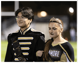 A person wearing glasses and a black and gold marching band uniform is standing outdoors with a slight smile. Description generated by AI